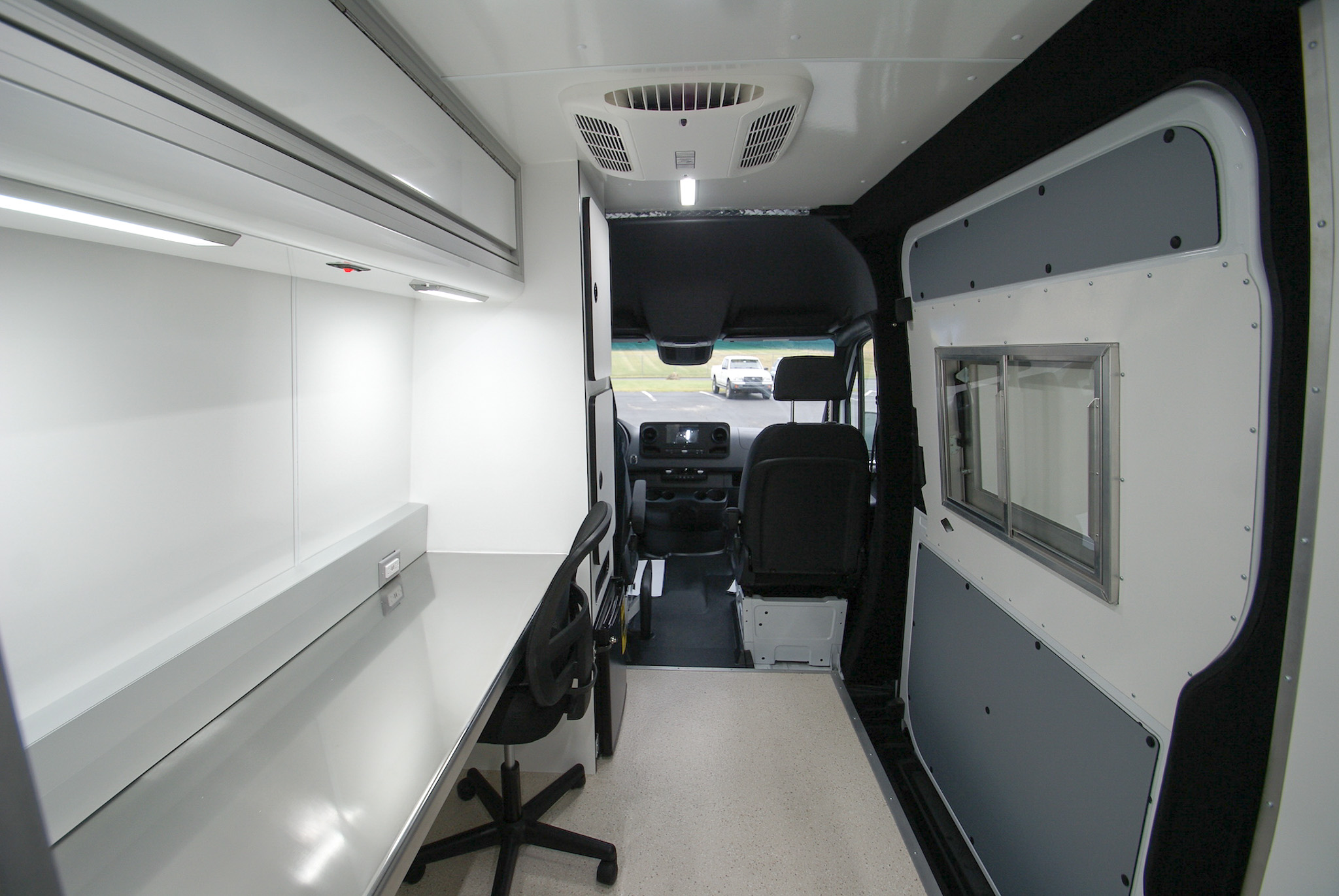 A direct view of the Mobile Immunization sprinter van's interior from the back room. There's a task chair near an open workspace, overhead cabinets, and both the driver and passenger seats up front.