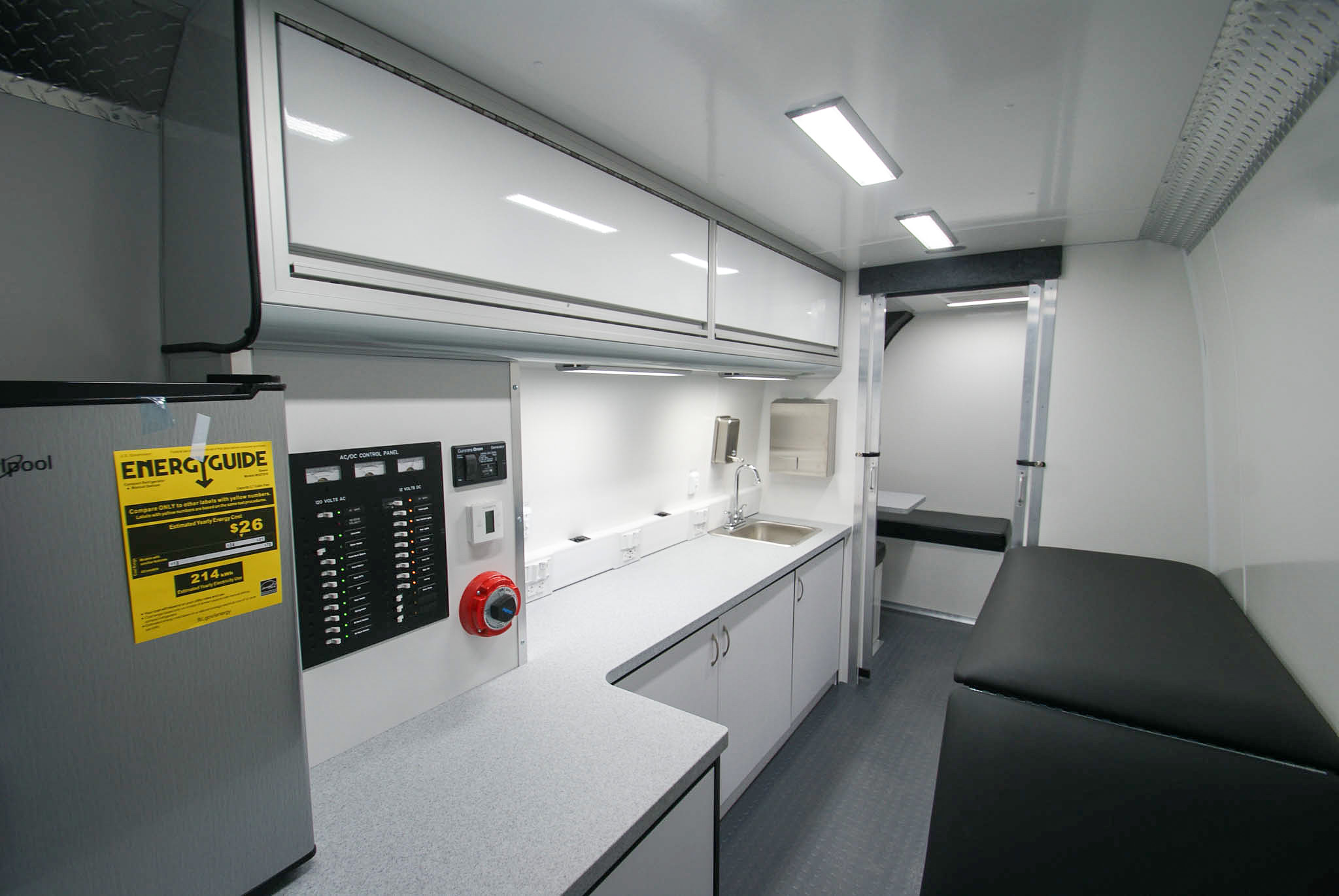 An angled view of the Mobile Medical Exam sprinter's back room as viewed from the rear entrance. You can see the refrigerator, electric control panel, countertops with a sink, overhead cabinets, and the exam bench. Past the open doors, you can see the waiting area.