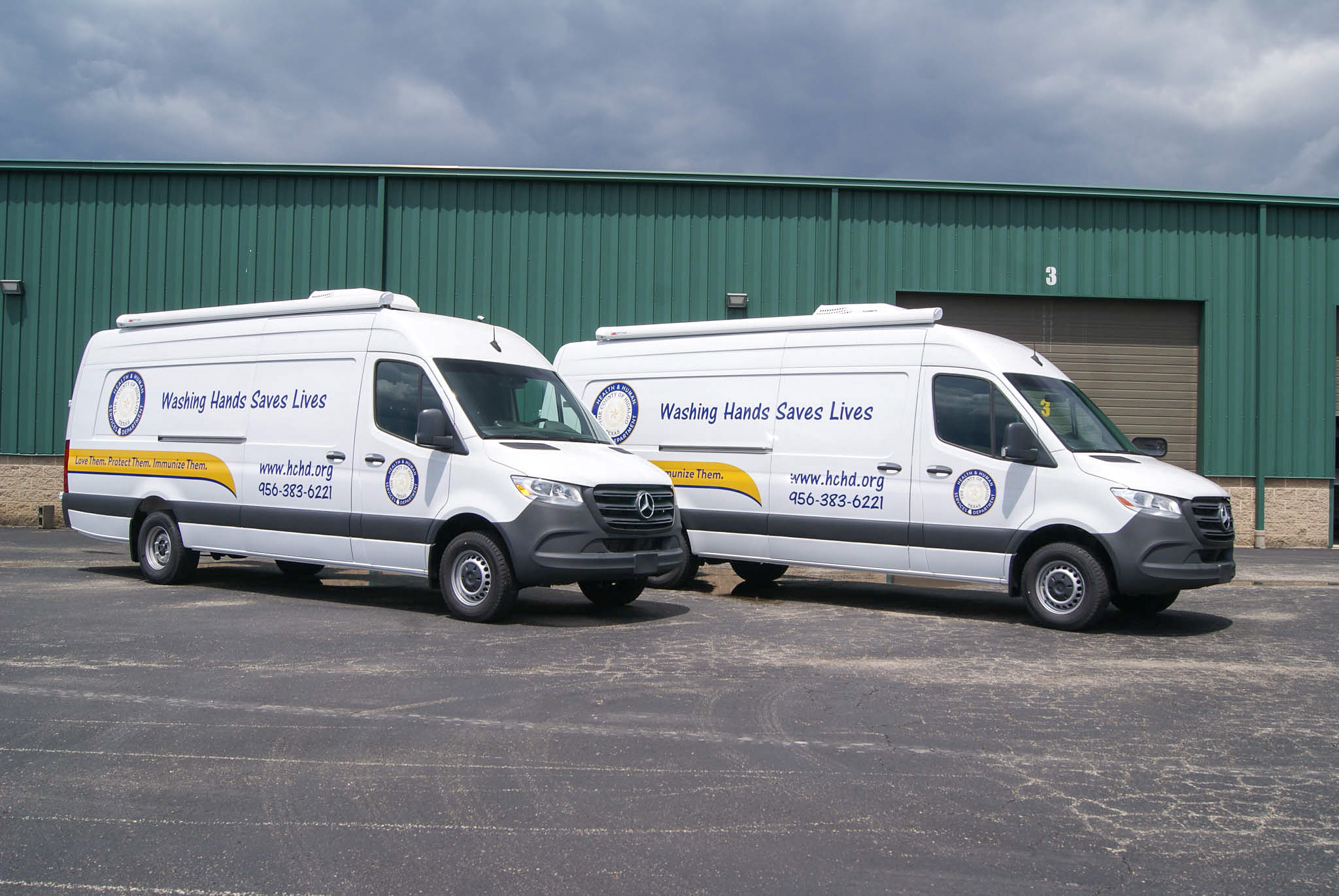 Two customized Mobile Medical Exam sprinter vans for Hidalgo County's Health Department are parked next to one another. The graphics feature their website, phone number, and the phrases, "Washing Hands Saves Lives," and "Love Them. Protect Them. Immunize Them."